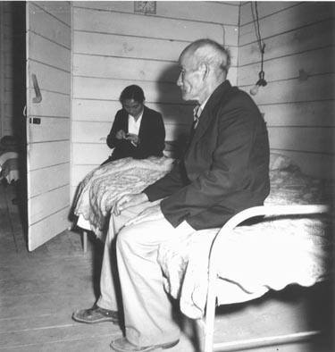 San Bruno Calif. June 16, 1942 "Old Mr. Konda in his barrack apartment, Tanforan Assembly Center, after supper.  He lives here with his two sons, his married daughter and her husband.  They share two small rooms together.  His daughter is seen behind him, knitting.  He has been a truck farmer and raised his family who are also farmers, in Centerville, Alameda County, where his children were born"