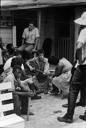 After giving a concert in a cotton field in Greenwood, Bob Dylan plays behind the SNCC office. Bernice Reagon, one of the original Freedom Singers and today leader of Sweet Honey in the Rock, listens. Mendy Samstein sits behind Dylan and talks to Willie Blue